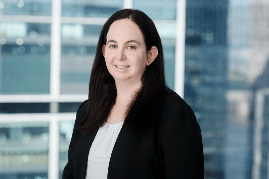Short white woman wearing a black blazer and white shirt in front of a background of windows.