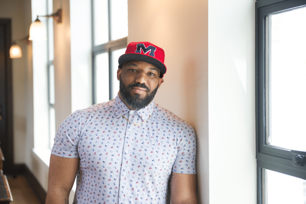 A Black man in a patterned shirt and hat stands against a wall.