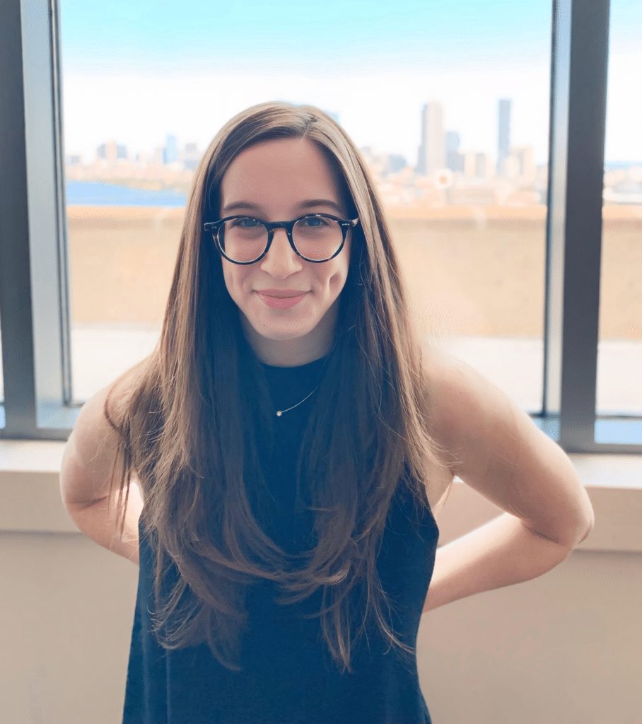 A woman with white skin and long brown hair and dark rimmed round glasses smiles in front of a window. She is wearing a black sleeveless shirt.