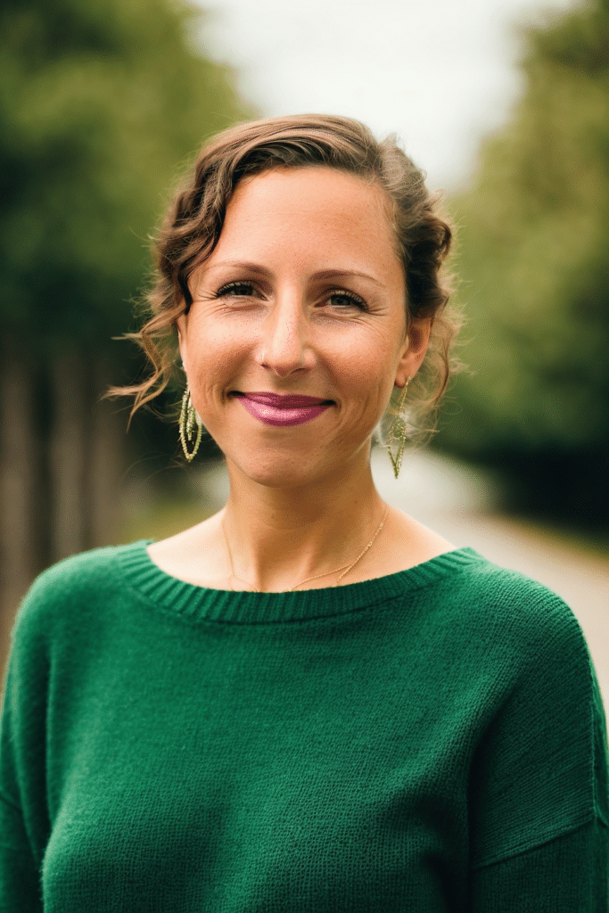 A smiling, light-skinned woman with curly brown hair that is pulled back, pink lipstick, and big round earrings. She is wearing a green sweater and stands against a blurred green backdrop of trees.