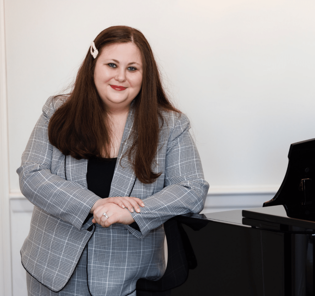 A white woman with long, dark hair in a patterned skirt suit smiles, leaning on a grand piano.