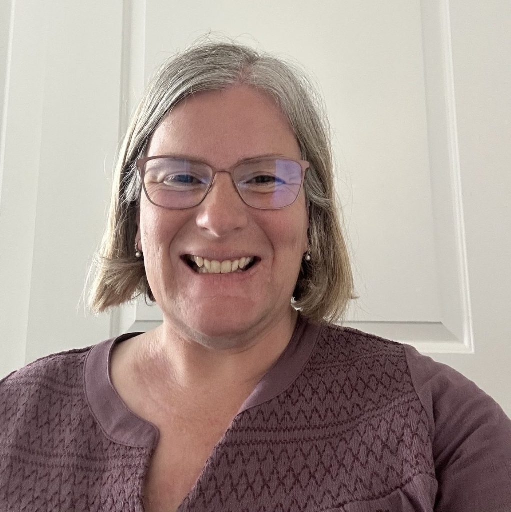 A white woman with salt and pepper hair wearing peach rimmed glasses and a purple peasant top against a white background.
